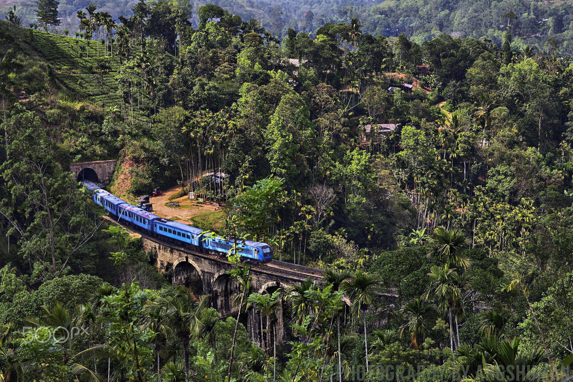 A blue train crossing the Nine Arch Bridge near Ella, Sri Lanka, a stone viaduct of nine arches set among dense green hillside vegetation.