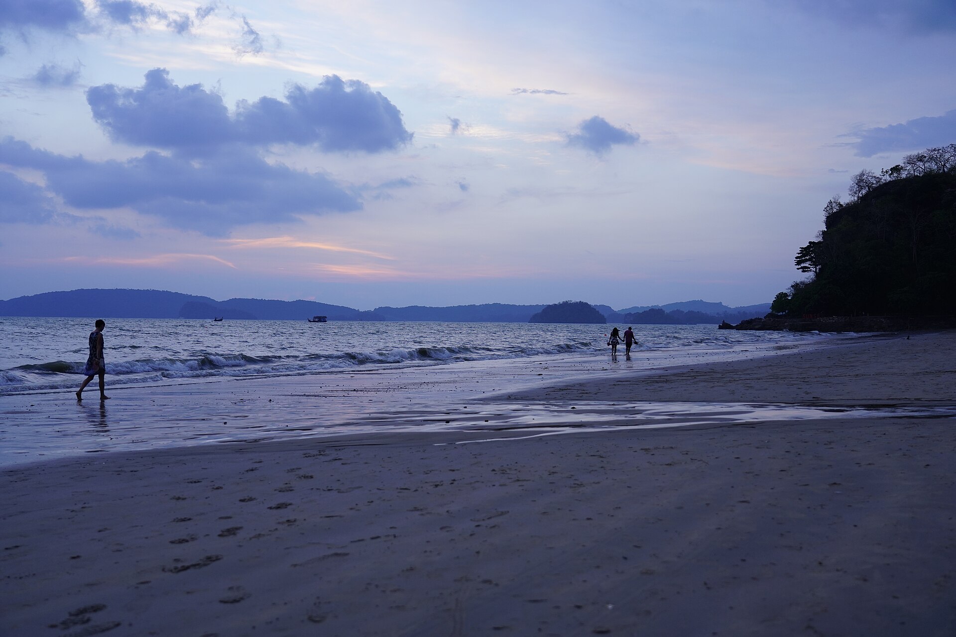 Ao Nang beach in Krabi, with longtail boats moored in shallow green water below forested limestone headlands.