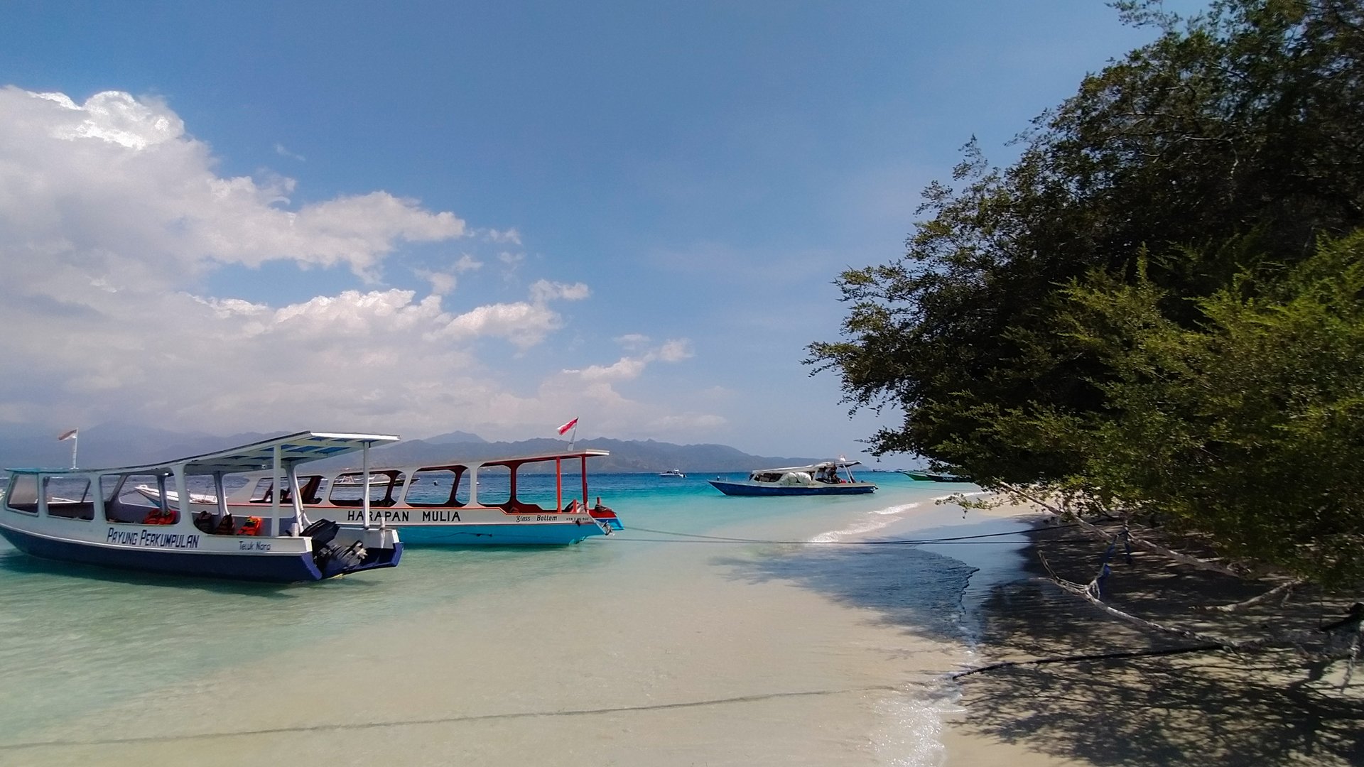 Wooden ferry boats moored on the shore at Gili Trawangan, with clear turquoise water and Gili Meno visible on the horizon.
