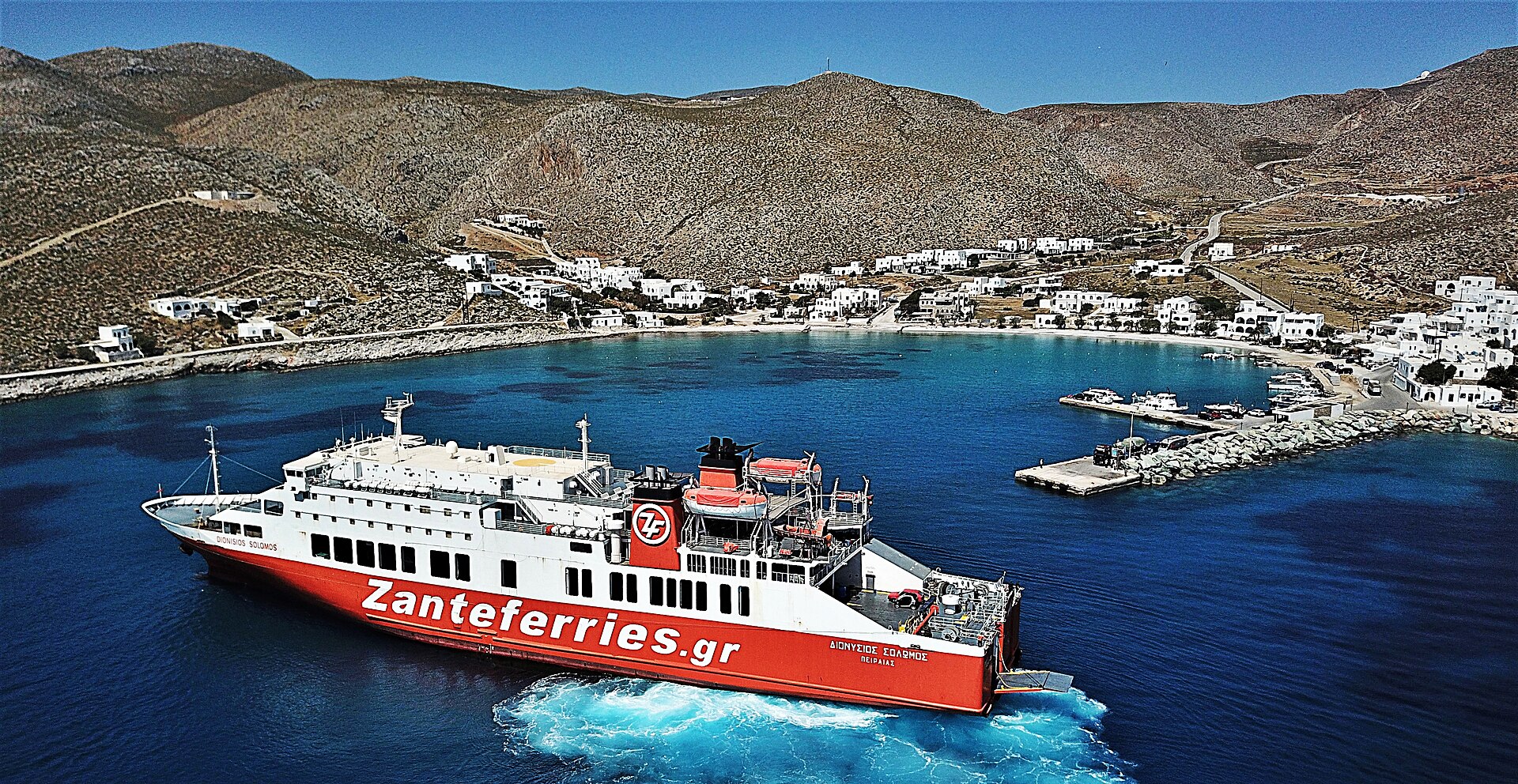 A blue and white Greek car ferry arriving at the small harbour of Folegandros in the Cyclades, with bare hillsides rising behind the quay.