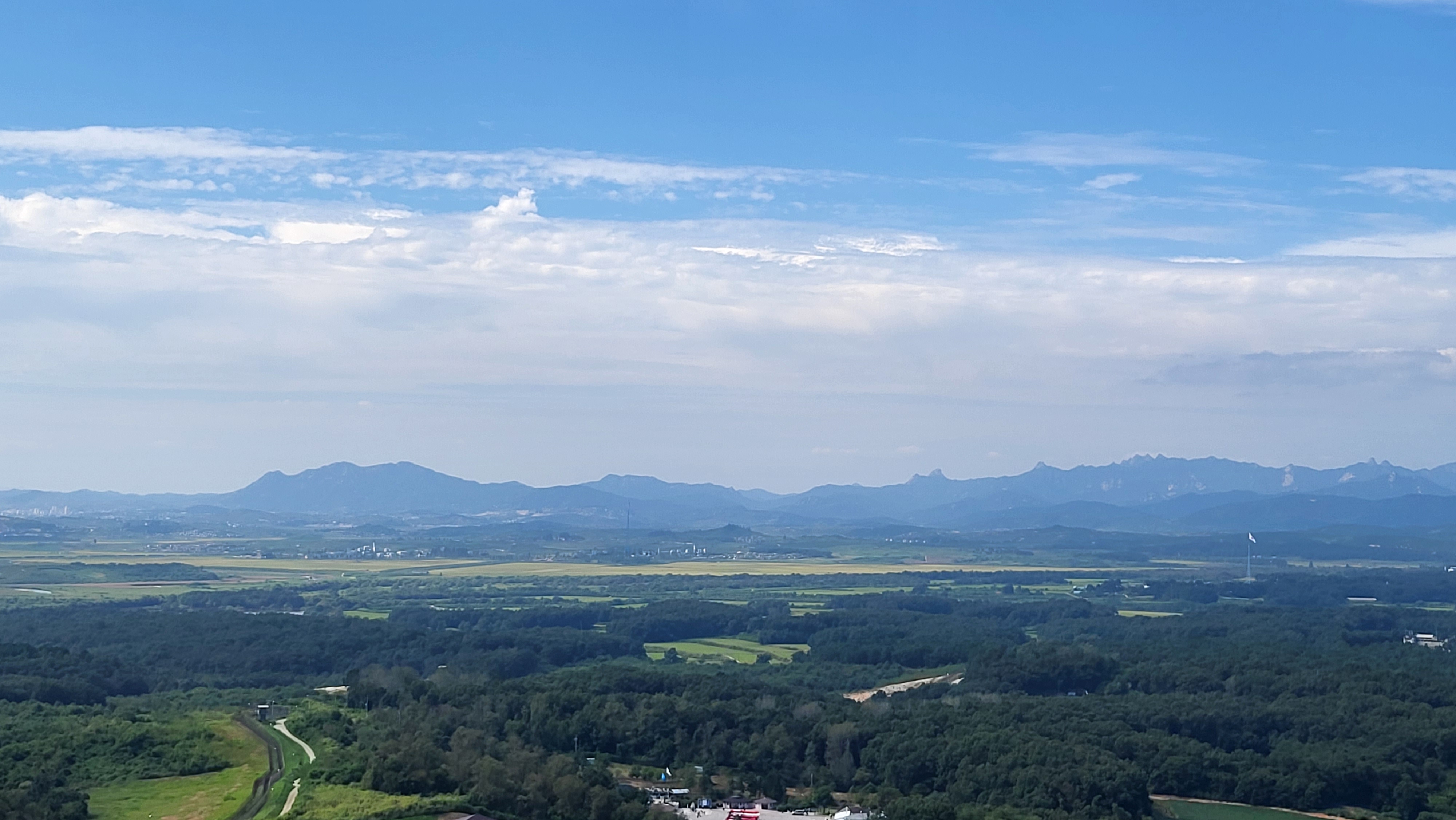 A wide view from the Dora Observatory over forest and farmland in the Demilitarized Zone toward distant hills in North Korea, under a blue sky with scattered cloud.