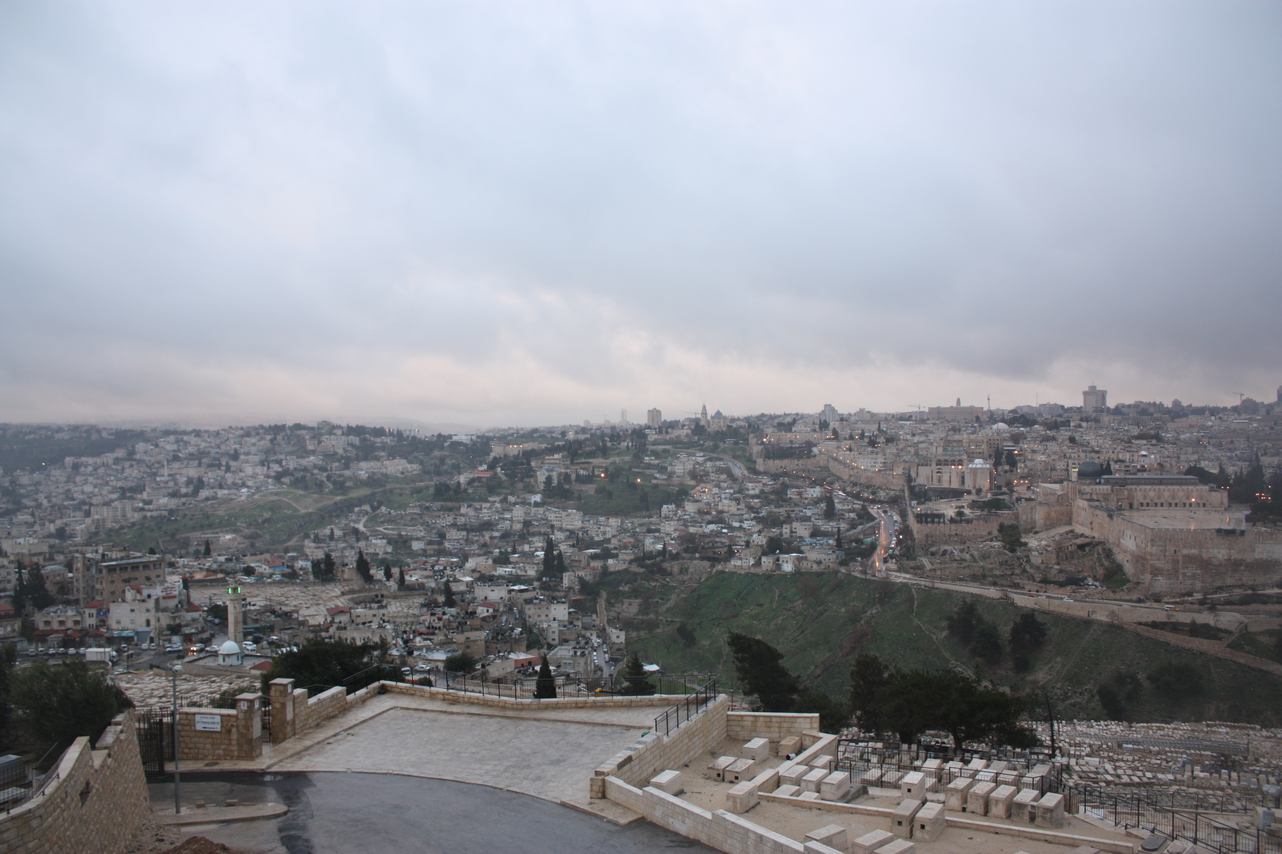 A hazy view across East Jerusalem from the Mount of Olives, with the Old City walls and the Dome of the Rock visible on the far ridge.