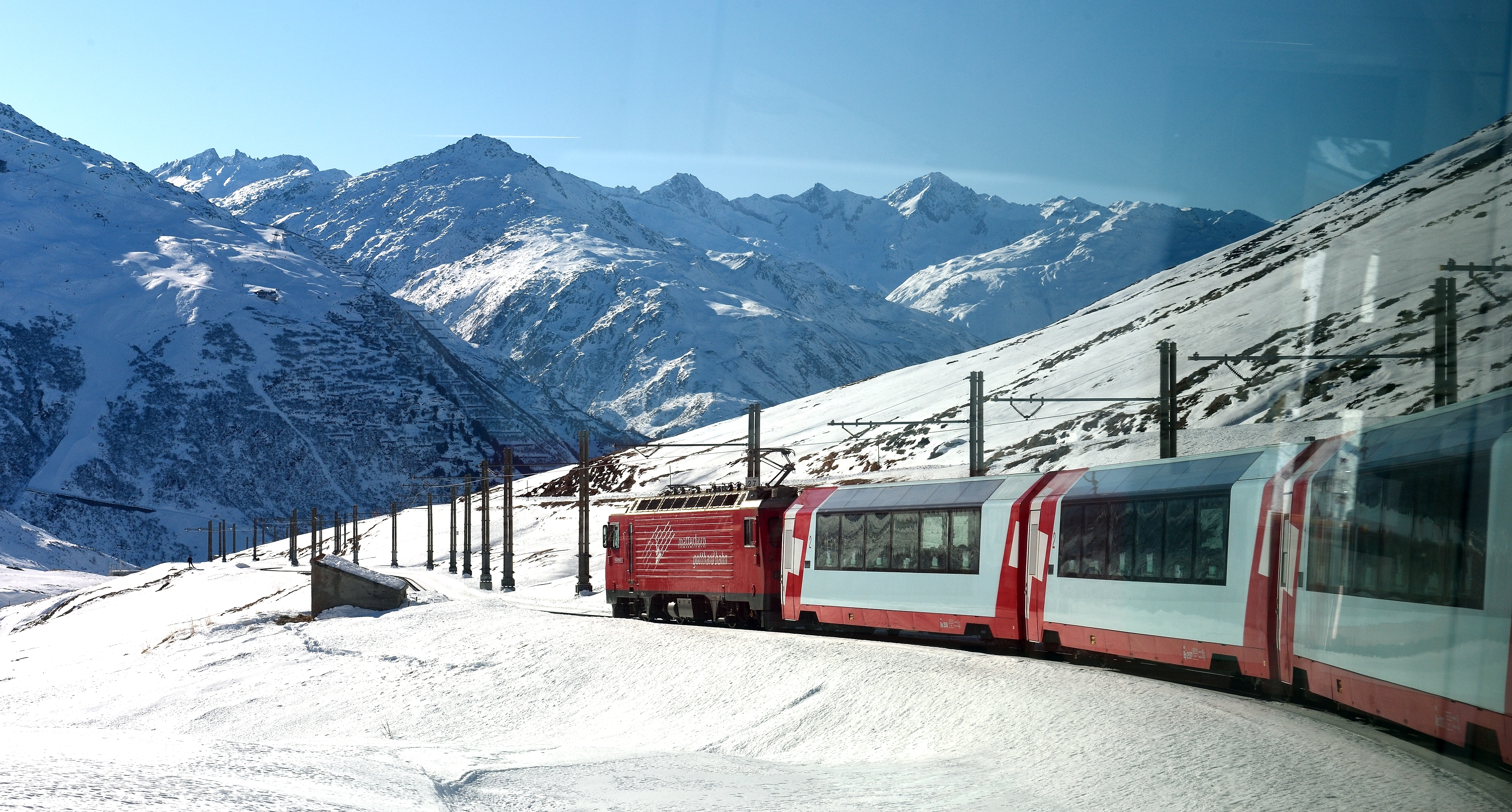 The Glacier Express crossing a viaduct east of Andermatt in the Swiss Alps, red carriages against steep grey mountainsides on the route to Zermatt.