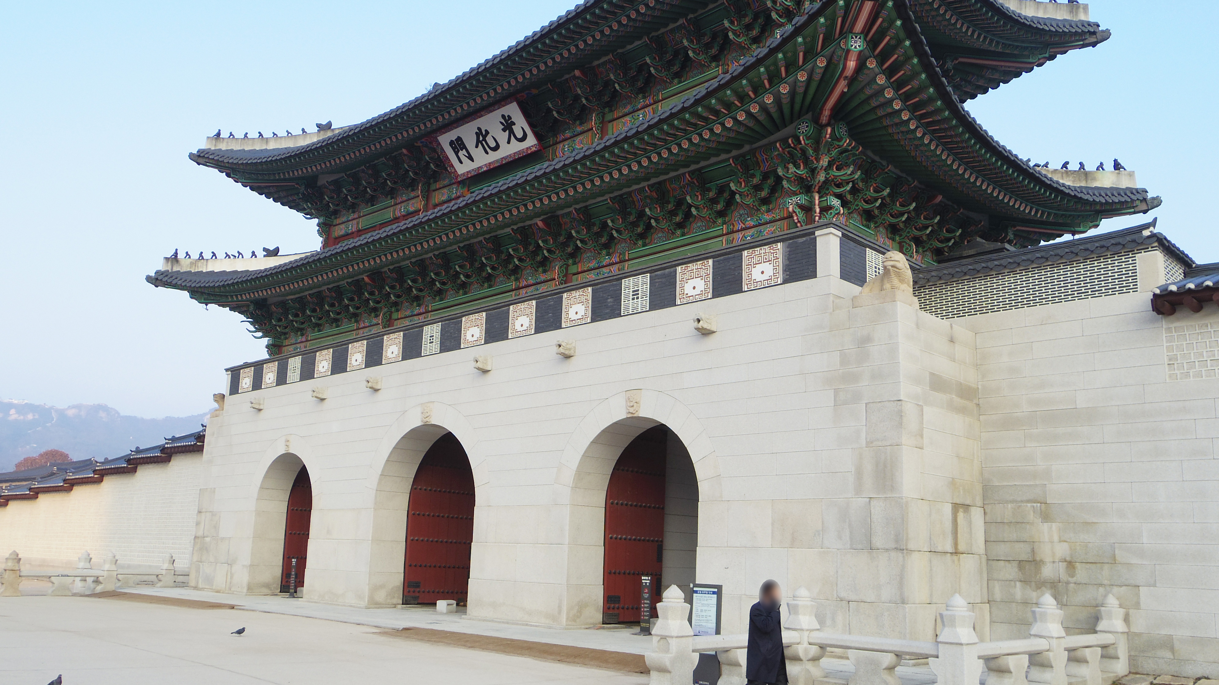Gwanghwamun, the main southern gate of Gyeongbokgung Palace, a three-arched stone base with red doors beneath a two-tier green and gold painted wooden roof.