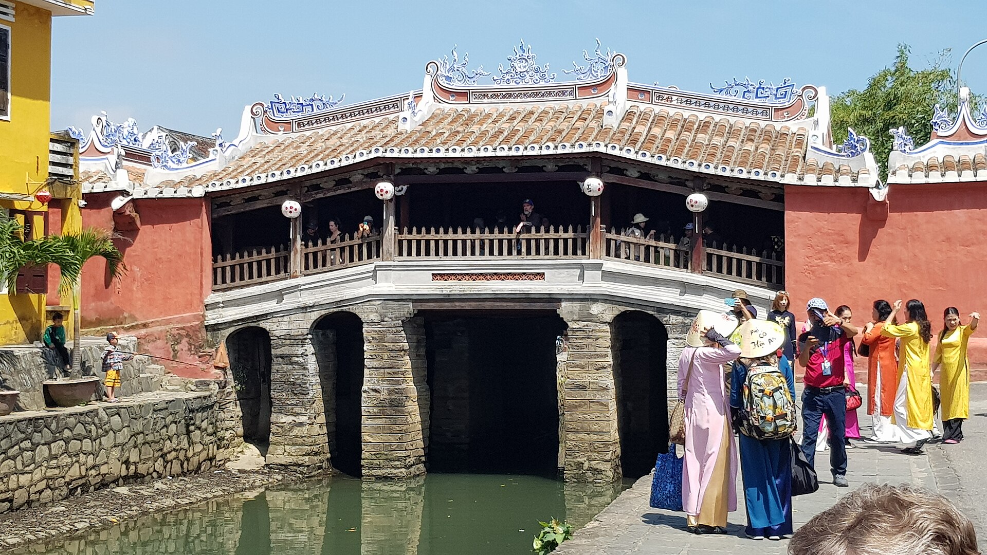 The Japanese Covered Bridge in Hoi An, a tiled roof over a stone arch across a narrow canal, with visitors gathered on the walkway beside it.