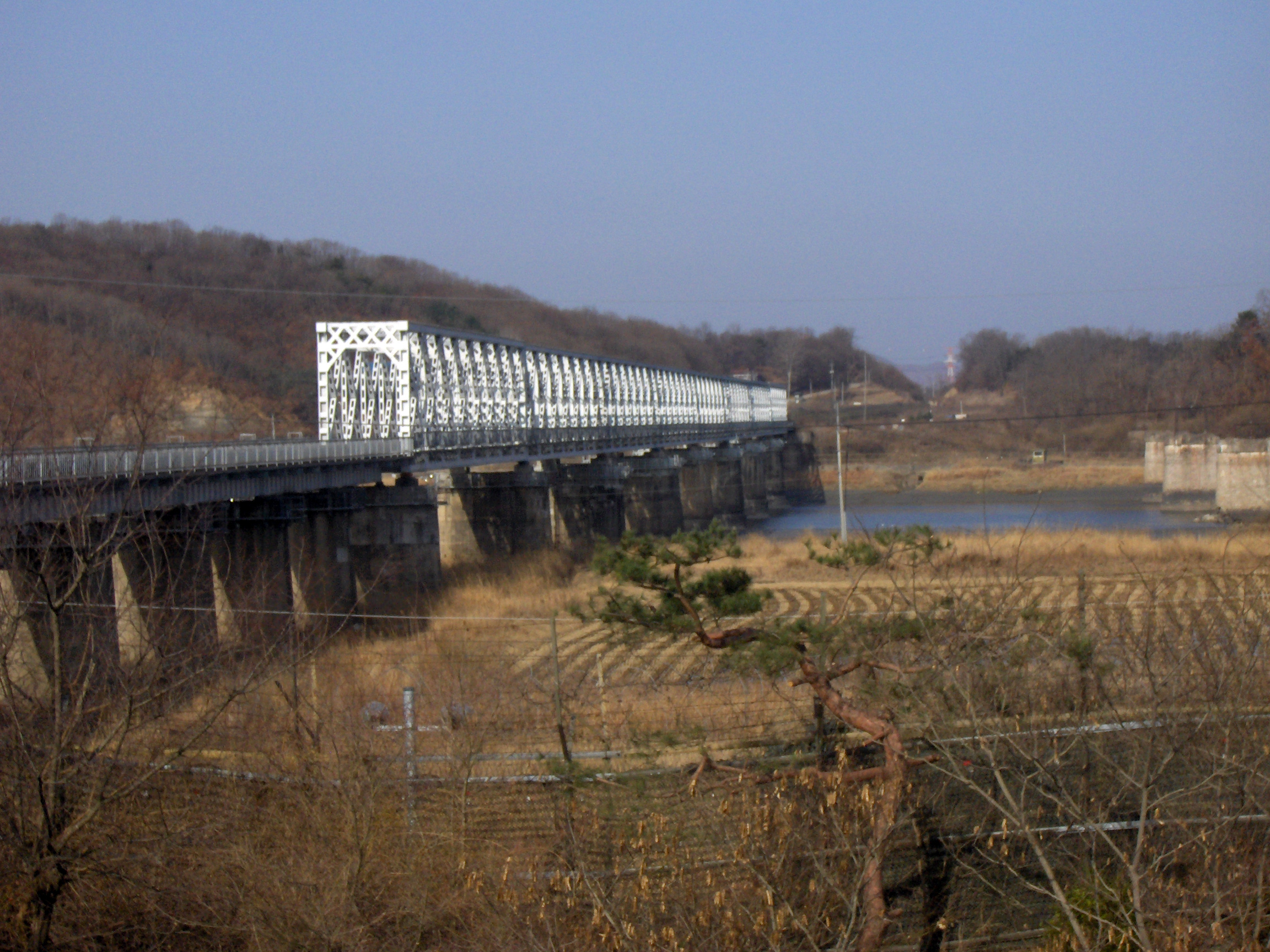 A long white steel truss railway bridge, the Freedom Bridge, crossing a river valley at Imjingak with barbed wire fencing in the foreground and bare winter hills behind.