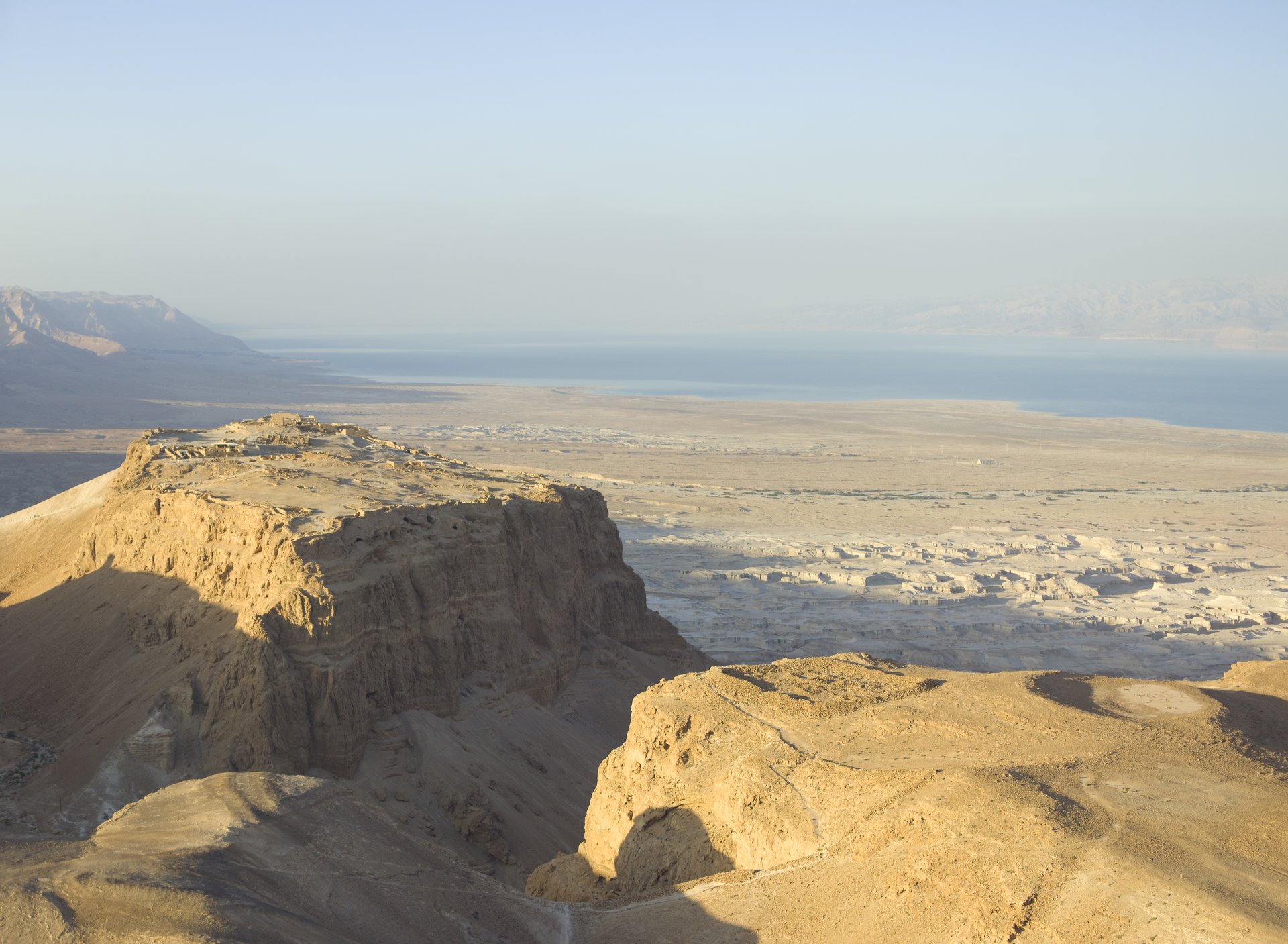 An aerial view of Masada's flat summit and sheer cliffs above the Dead Sea, with the ancient Roman siege ramp and desert visible below.