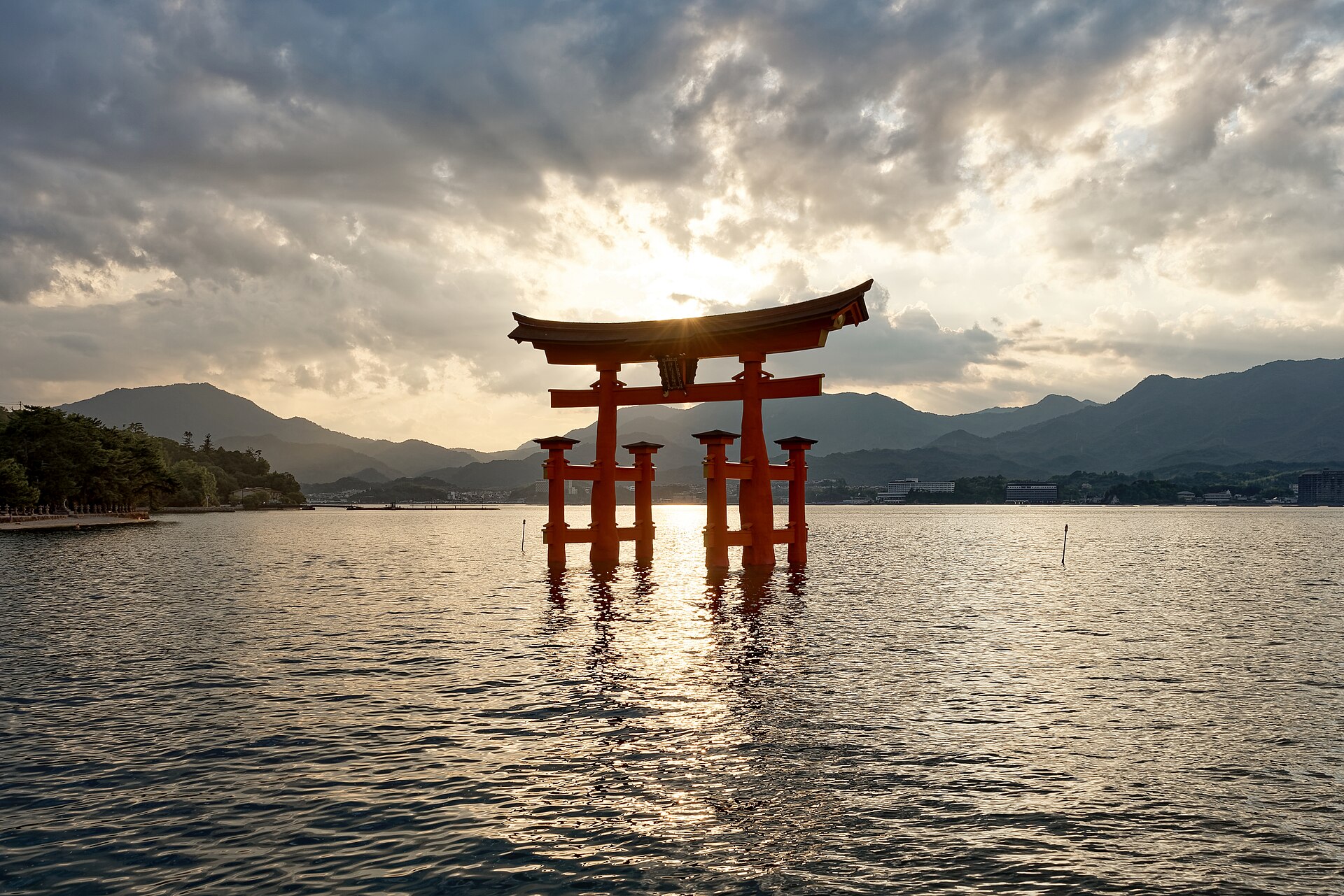 The vermilion floating torii gate of Itsukushima Shrine standing in shallow water at sunset, with wooded hills behind.