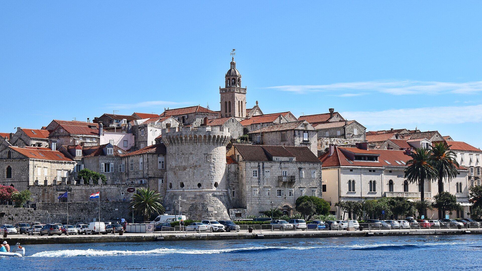 The walled old town of Korčula on its small peninsula, red roofs and a bell tower surrounded by clear sea and wooded hills.