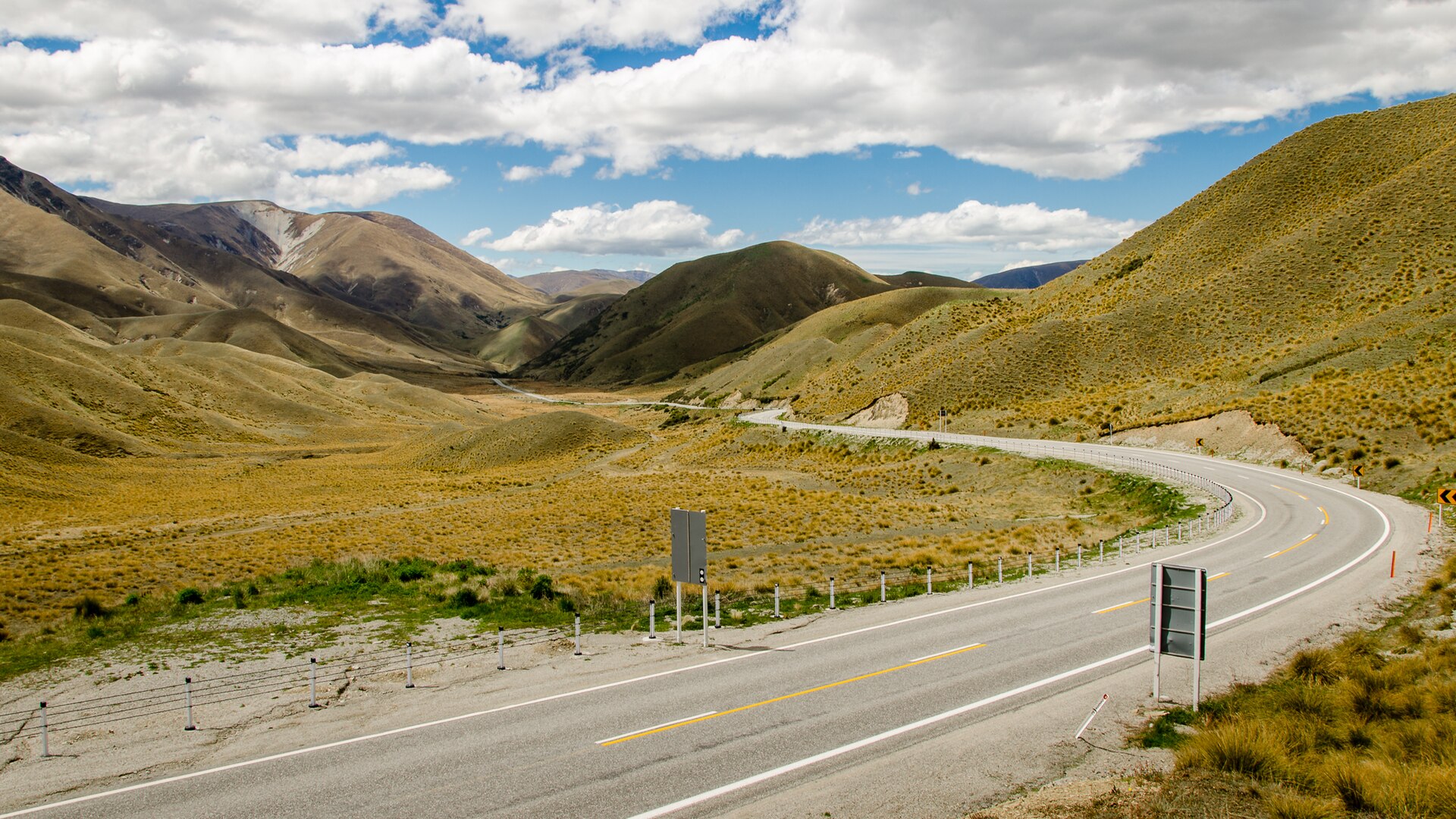 The tussock-covered hills of the Lindis Pass in Otago, New Zealand, with State Highway 8 curving through the valley under a clear sky.