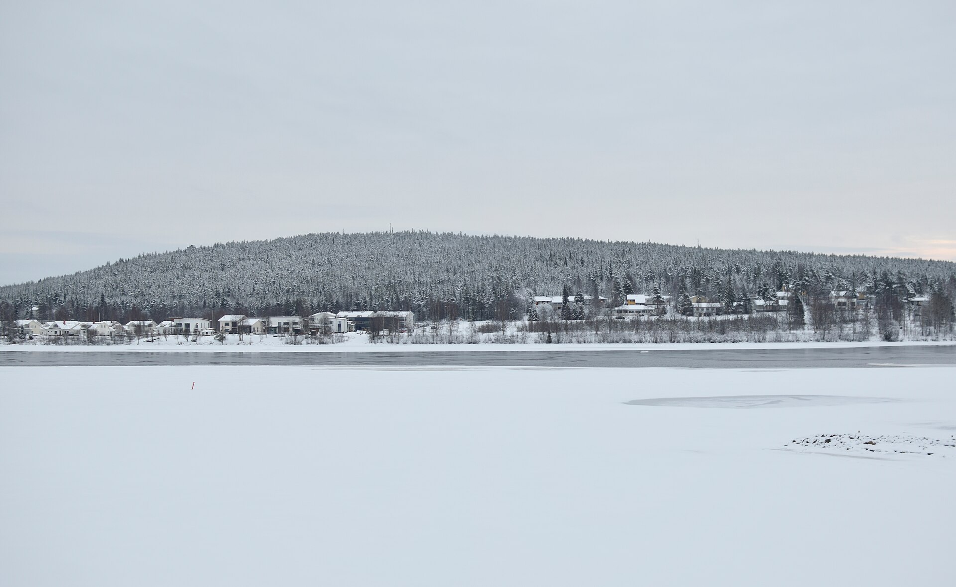 A forested hill rising behind a few houses across a frozen, snow-covered river under a pale grey winter sky.