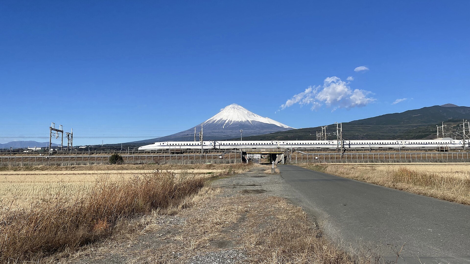 A white and blue Shinkansen train crossing open ground in Shizuoka with the snow-capped cone of Mount Fuji behind it.