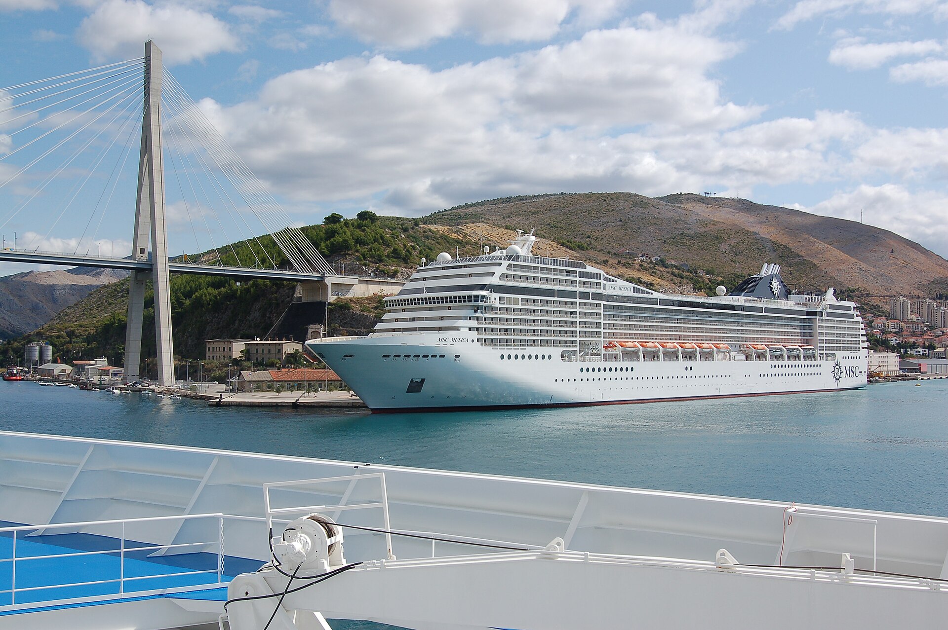 A large white cruise ship tied up at the quay at Gruž, the commercial port that serves Dubrovnik, with hills rising behind the harbour.