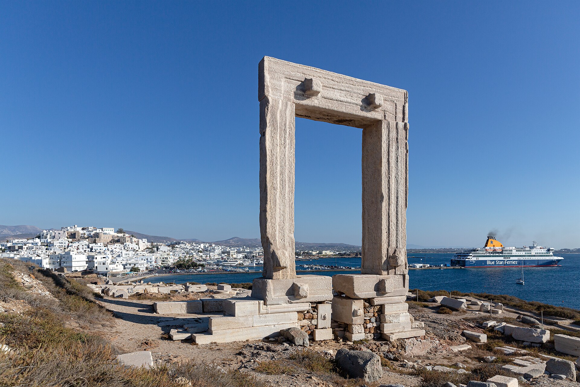 The Portara stone doorway on the islet at Naxos town, framing a Blue Star ferry in the harbour beyond it.