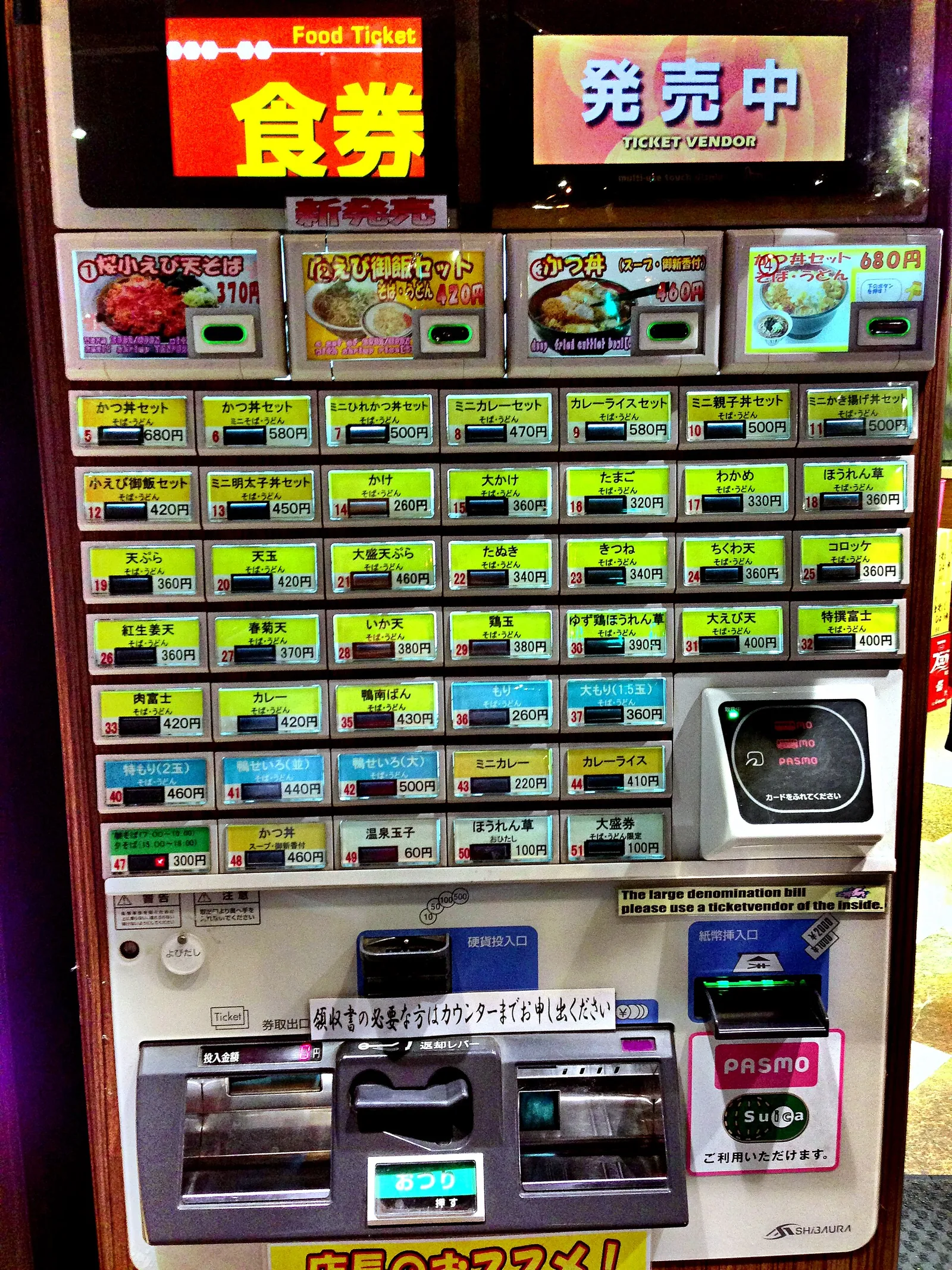 A food ticket vending machine outside a Fuji-soba shop in Akihabara, with rows of numbered buttons listing dishes and prices in yen, and a PASMO and Suica card reader on the right.