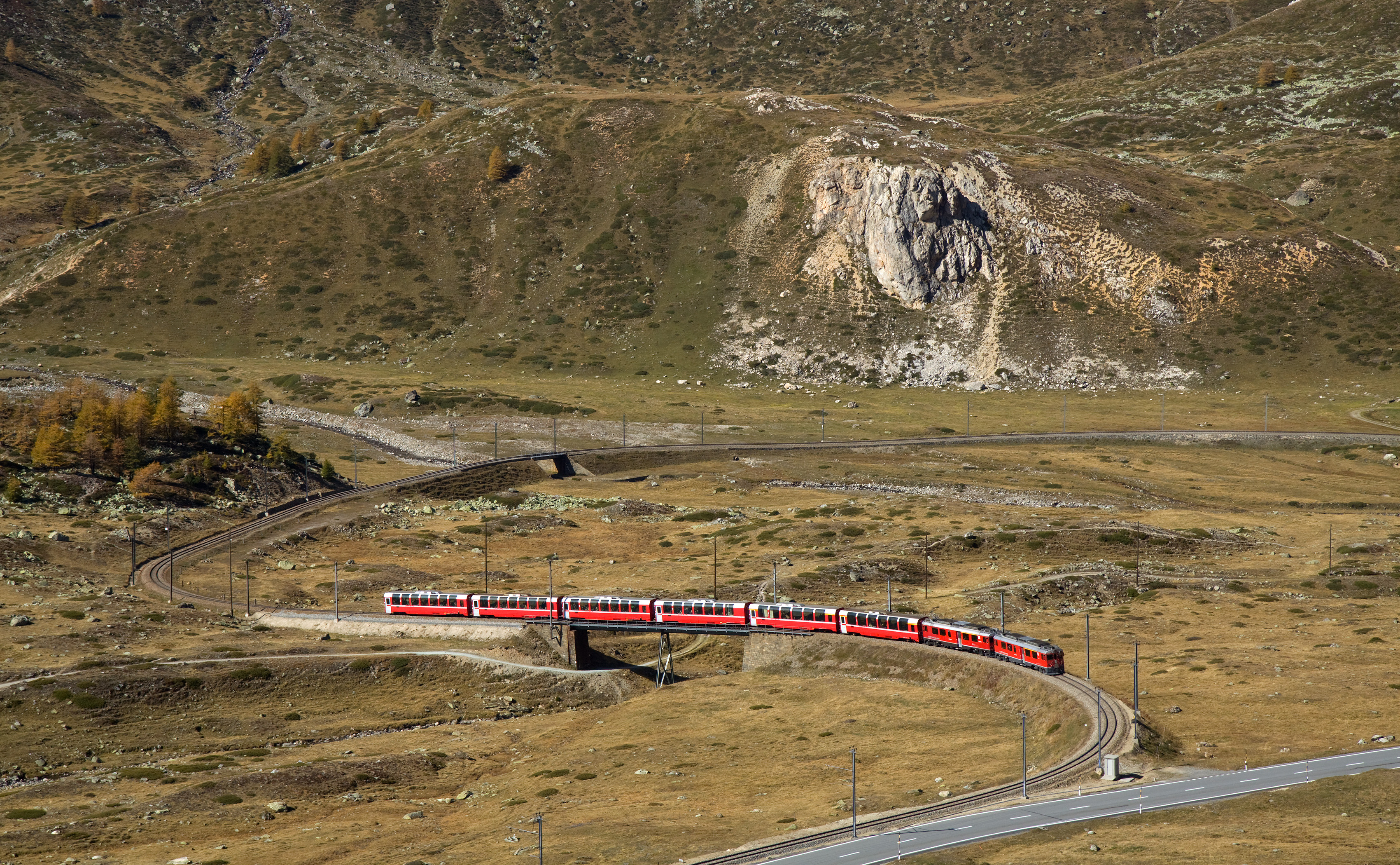 The Bernina Express climbing a steep grade toward Ospizio Bernina, the train crossing a stone bridge with bare alpine slopes in the background.