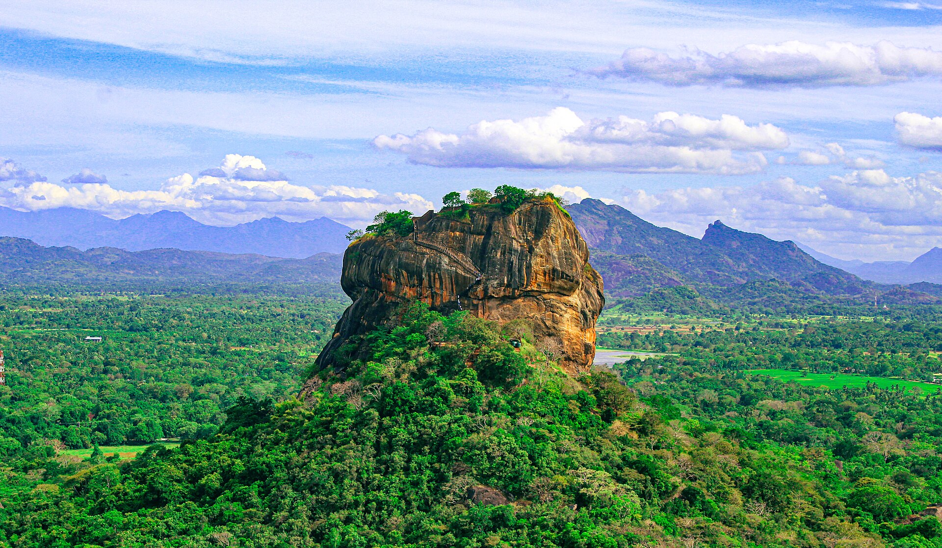 The rock fortress of Sigiriya rising out of flat green forest in central Sri Lanka, with cloud building above it.