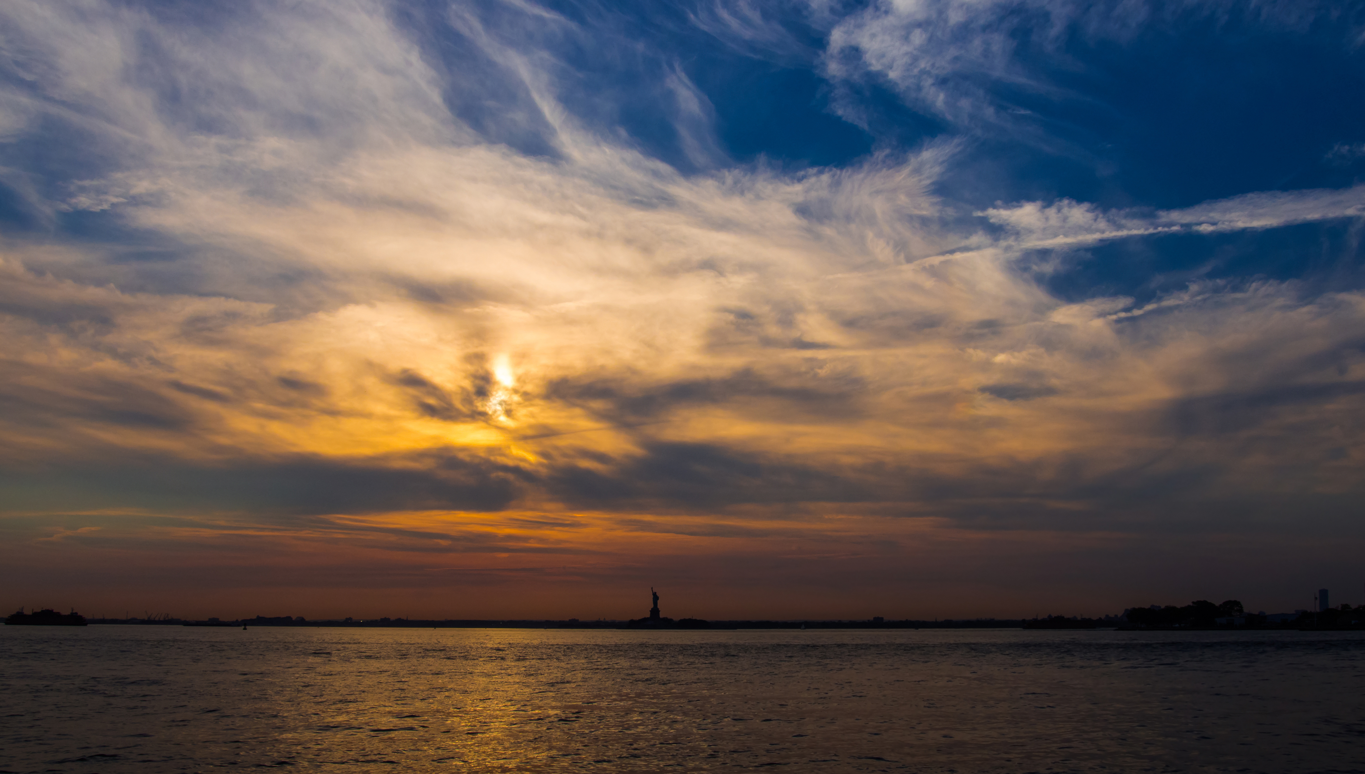 The Statue of Liberty seen across Upper New York Bay at dusk, with the Manhattan skyline faint on the horizon behind it.