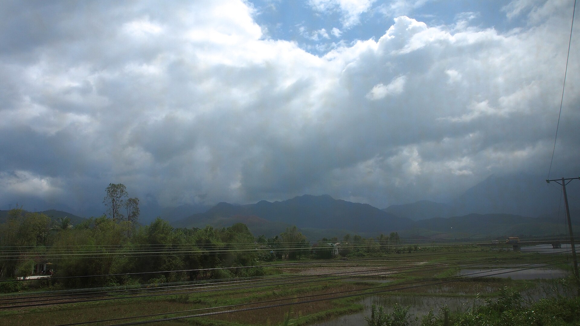 Railway tracks running through green paddy fields towards a range of mountains in central Vietnam, under a heavy bank of cloud.