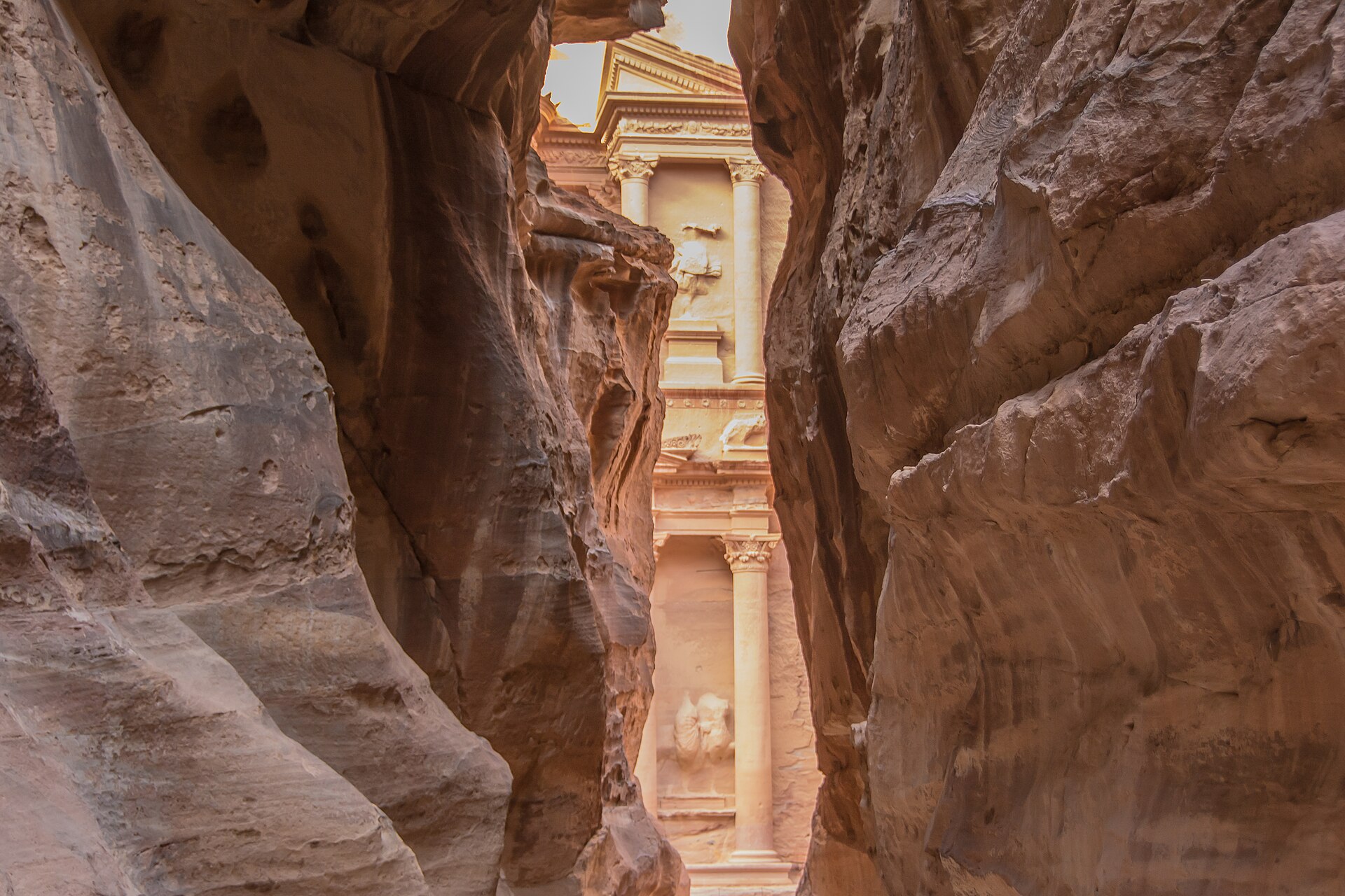 The Treasury at Petra seen through the narrow opening at the end of the Siq gorge, its carved facade lit while the rock walls in the foreground stay in shadow.