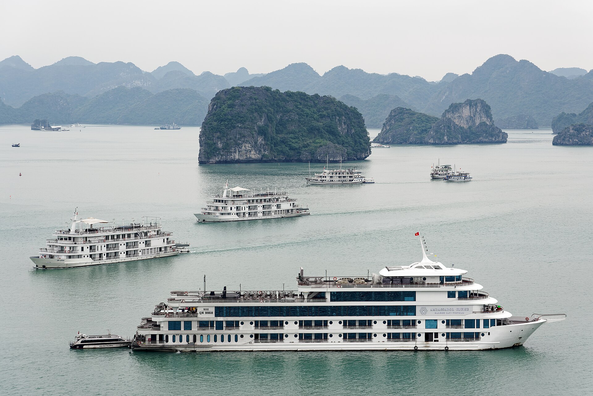 Cruise boats moving between forested limestone karst islands in Ha Long Bay, Vietnam, under a pale hazy sky.