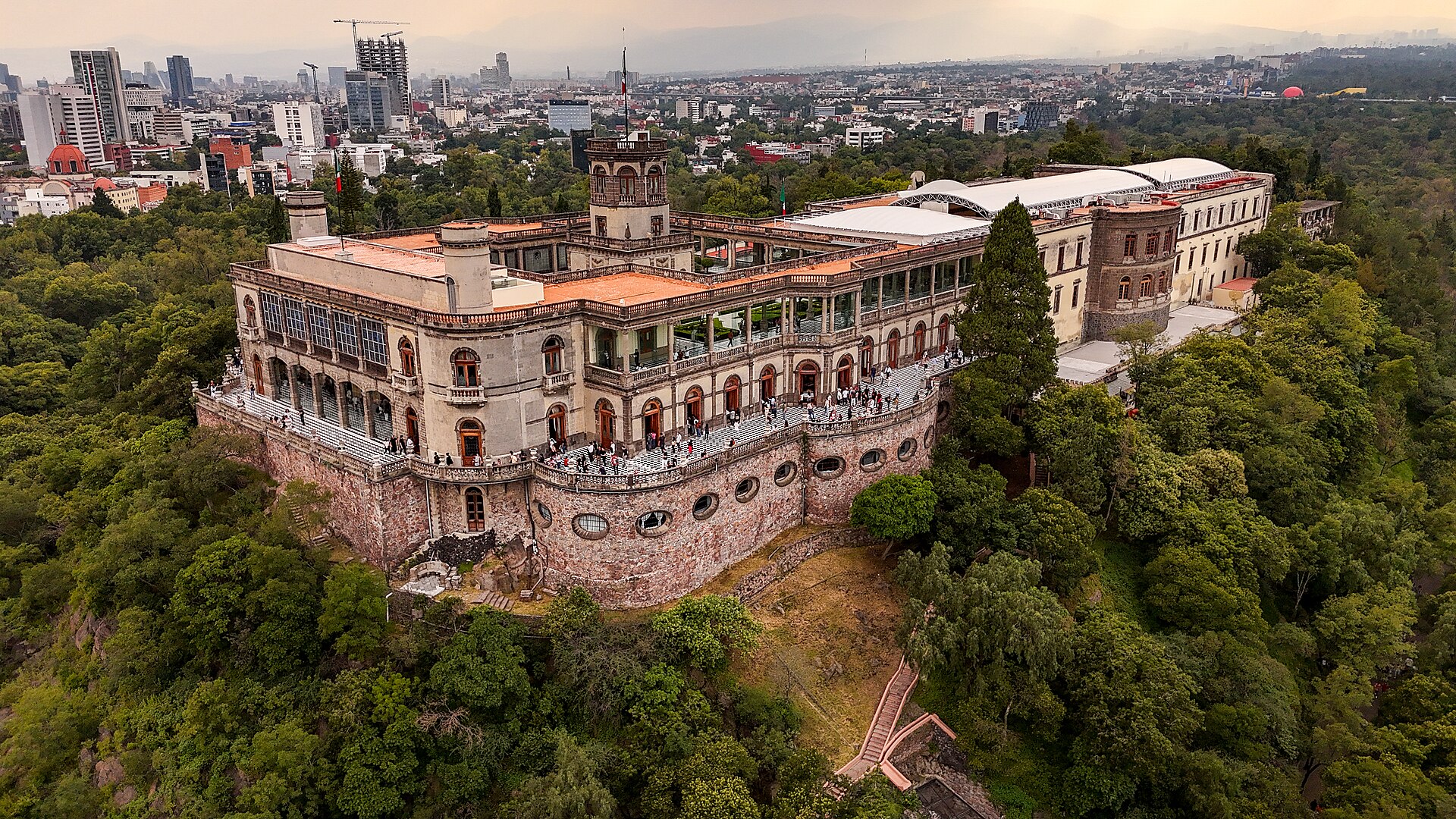 An aerial view of Chapultepec Castle standing on its wooded hill above Mexico City, with parkland and city buildings spreading out behind it.