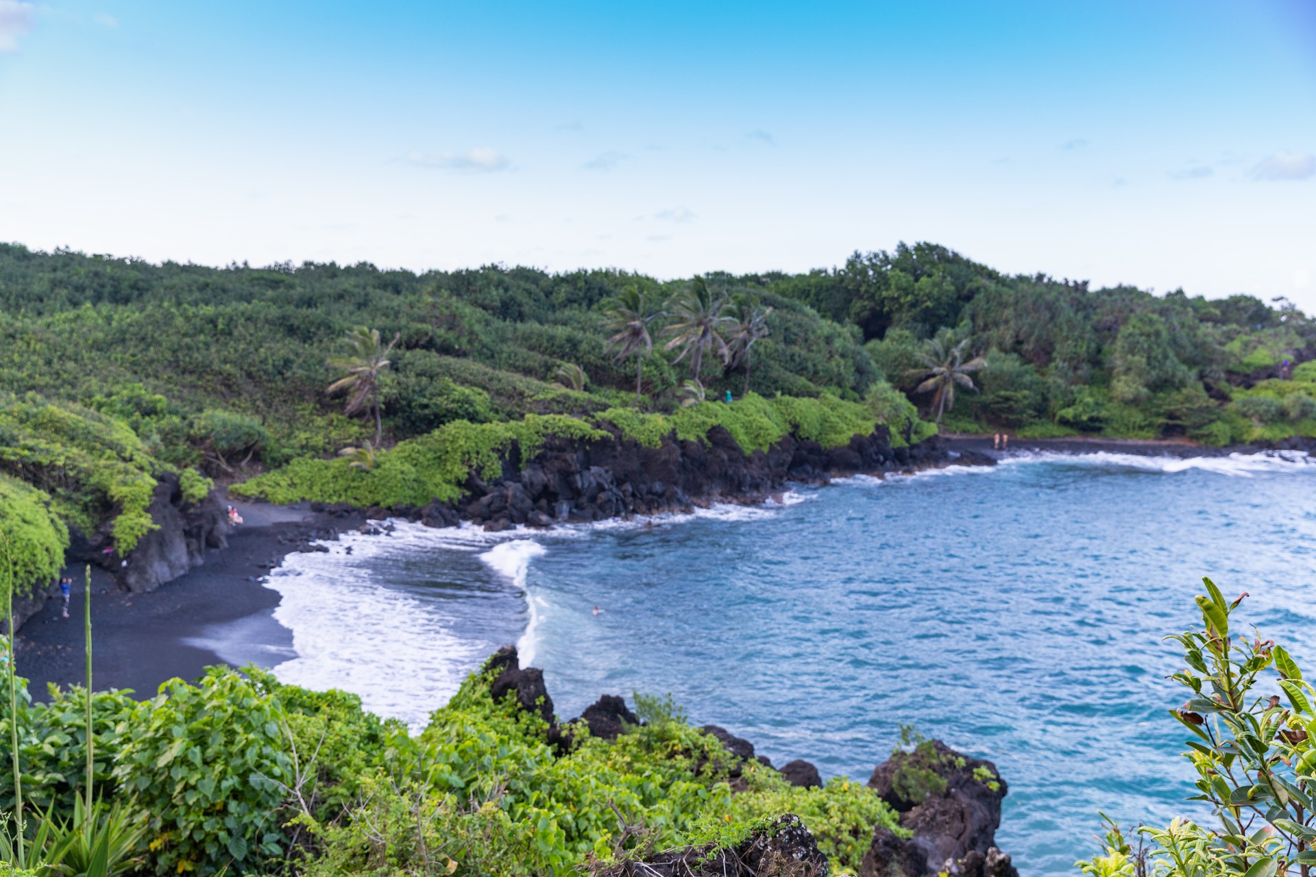 Waiʻānapanapa's black sand beach on Maui's north shore, dark volcanic sand meeting turquoise water beneath green cliffs.